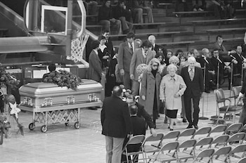 Ataúd metálico con arreglo floral en un gimnasio. Varias personas, algunas con gafas, caminan frente a sillas plegables. Gradas y canasta de baloncesto visibles al fondo