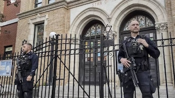 Dos policías con equipo táctico y rifles custodian un edificio de ladrillo con puertas arqueadas y una valla. Se ven estrellas de David en la fachada