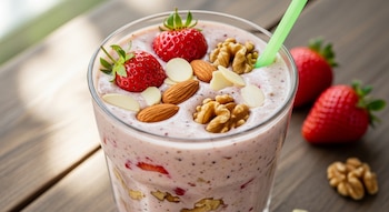Primer plano de un batido de fresas, almendras y nueces en un vaso, con un sorbete verde. Fresas y frutos secos adornan la superficie y la mesa.