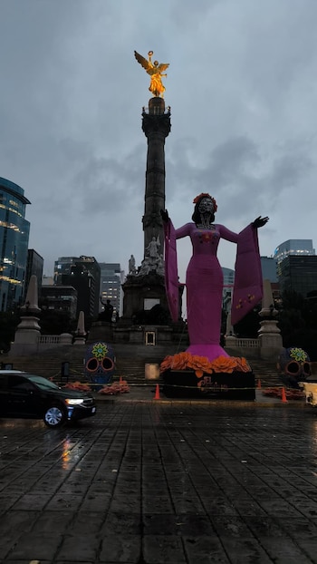 Escultura gigante de una Catrina rosa frente al monumento del Ángel de la Independencia sobre una avenida mojada con edificios al fondo