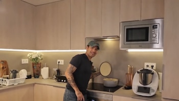 Hombre sonriente con gorra y tatuajes en la cocina. Sostiene la tapa de una olla sobre una estufa. Hay un microondas, gabinetes y utensilios