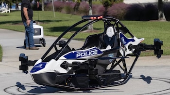 Vehículo aéreo monoplaza con hélices y rotulación policial sobre pavimento, con una persona y un pequeño robot de reparto al fondo