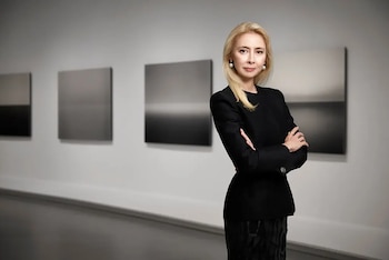 Melissa Chiu, una mujer rubia de mediana edad con un traje negro, de pie con los brazos cruzados en una galería de arte frente a pinturas abstractas en tonos grises