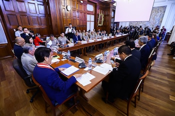 Numerosos funcionarios y expertos sentados alrededor de una gran mesa de madera en una sala de reuniones formal con paneles de madera y una pantalla de proyección