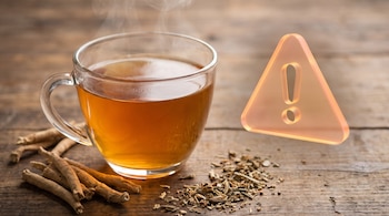Una taza de cristal con infusión humeante, raíces y trozos de ashwagandha seca sobre una mesa de madera, junto a un signo de advertencia triangular.