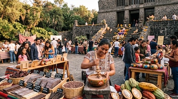 Una mujer bate cacao en un cuenco junto a mesas con barras de chocolate, mazorcas y frutos en un patio con edificio de piedra al fondo.