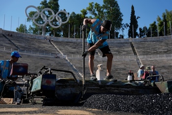 Dos hombres trabajan con una máquina y palas aplicando material negro en una superficie, con un estadio de gradas y anillos olímpicos al fondo