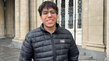 Primer plano de un joven con anteojos y chaqueta negra acolchada con el logo Fundación YPF. Fondo de edificio con columnas y puerta principal con número 609