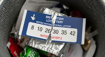 Billete de la Lotería Nacional británica roto con números 8, 10, 26, 30, 35 y 42 en el fondo de un cubo de basura con residuos.