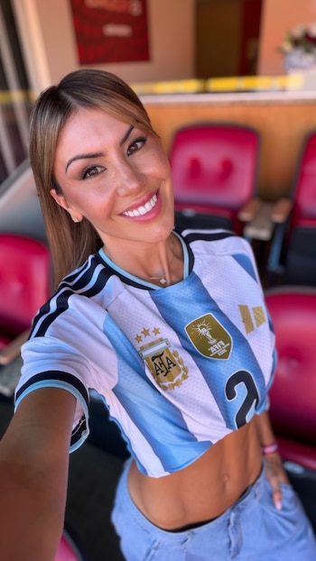 Mujer de cabello castaño claro y ojos marrones sonríe en primer plano con una camiseta de fútbol de Argentina con tres estrellas y jeans, en un estadio con asientos rojos