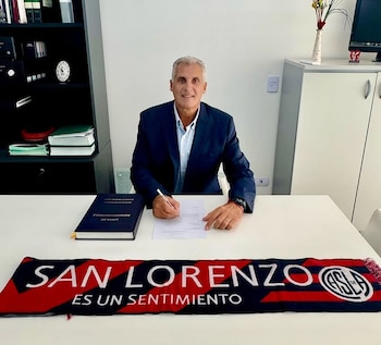 Alberto Acosta, un hombre de cabello gris y traje azul, sentado en un escritorio blanco, firmando un documento con una bufanda de San Lorenzo
