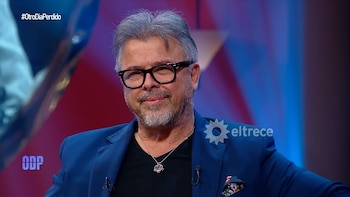 Primer plano de Donato De Santis, un hombre de cabello canoso, barba, gafas de montura negra y blazer azul, sonriendo sutilmente frente a un fondo difuso