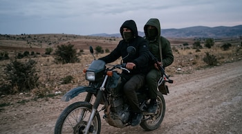 Dos hombres con capucha y el rostro cubierto viajan en una motocicleta por un camino de tierra, y el pasajero lleva un fusil.