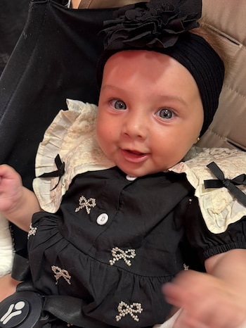 Um bebê com vestido preto com laços, gola branca com renda e laços pretos e chapéu preto com flores. A criança tem olhos azuis e pele clara.