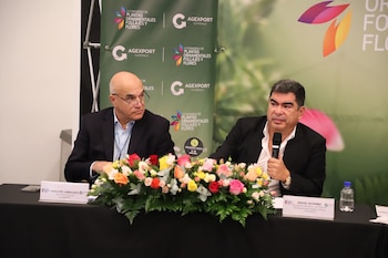 Dos hombres en una mesa con arreglo floral. Un hombre calvo con gafas mira a su derecha. Otro hombre sostiene un micrófono. Un banner verde de AGEXPORT en el fondo