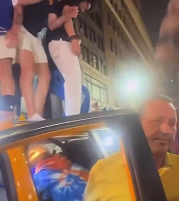 Varias personas de pie sobre el techo de un vehículo amarillo. Un hombre sonriente con camisa amarilla en primer plano. Luces brillantes y edificios de noche al fondo