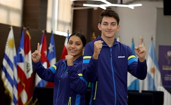 Un joven y una joven sonríen, visten chándales azules. El joven levanta el índice y la joven el puño. Banderas de Uruguay, Cuba y Argentina al fondo