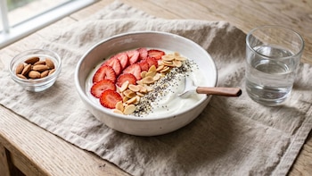 Tazón con yogur, rodajas de frutilla, almendras fileteadas y semillas, junto a un recipiente pequeño con almendras y un vaso de agua