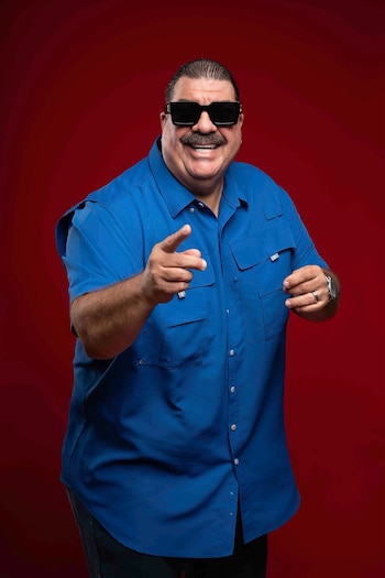 Hombre con bigote, gafas de sol negras, camisa azul de manga corta y pantalón oscuro. Apunta hacia adelante con la mano derecha. Usa reloj y anillo. Fondo rojo