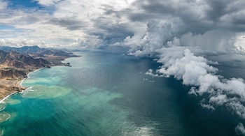 Vista aérea de una costa montañosa junto a aguas turquesas y una densa pared de nubes blancas sobre el océano Pacífico.