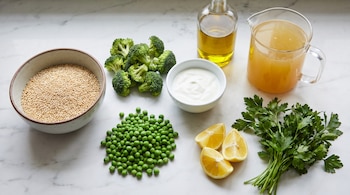 Quinoa en un cuenco, brócoli, arvejas, yogur, gajos de limón, perejil, una botella de aceite y una jarra de caldo sobre una mesada de mármol.