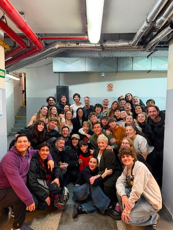 Grupo numeroso de personas sonrientes posa en un espacio interior con paredes blancas, tuberías rojas y conductos de ventilación metálicos. Hay un cartel de no fumar