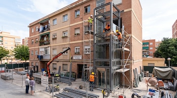 Varios obreros con cascos y chalecos trabajan en la estructura metálica de un ascensor sobre andamios junto a un edificio de ladrillo.