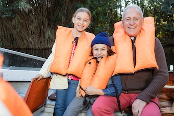 Un abuelo y sus dos nietas sonríen en un barco, usando chalecos salvavidas naranjas. La niña menor lleva un gorro azul. Fondo de vegetación frondosa