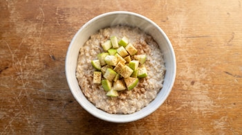 Vista cenital de un tazón blanco con avena cremosa, trozos de manzana verde y canela en polvo, sobre una mesa de madera bajo luz matutina natural.
