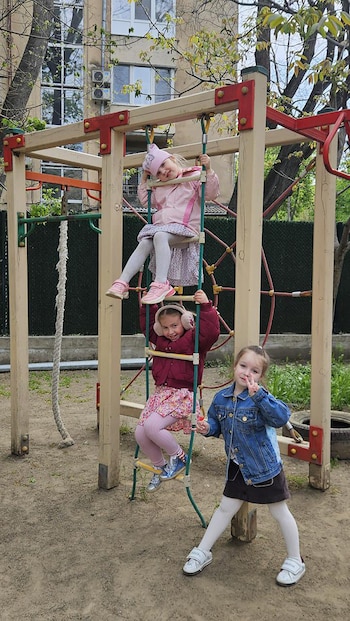 Tres niñas pequeñas juegan en un Playground. Dos escalan una estructura de cuerda y metal, mientras una tercera, vestida de mezclilla, posa con un signo de paz