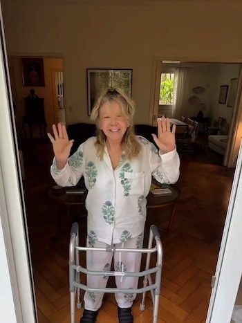 Una mujer en pijama blanco estampado permanece de pie con las manos levantadas detrás de un andador ortopédico en una habitación