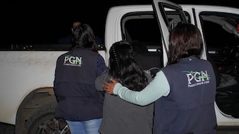 Tres personas vistas de espaldas, dos con chalecos de PGN, acompañan a una persona joven con cabello largo junto a una camioneta blanca embarrada en la oscuridad