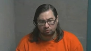 Primer plano de Armand Amar, un hombre con cabello oscuro y largo, gafas, barba y bigote, vistiendo una camiseta naranja