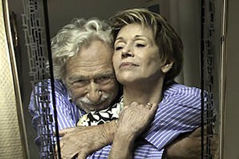 Un hombre y una mujer mayores abrazados. El hombre, de pelo gris y bigote, abraza a la mujer rubia de pelo corto. Ambos usan camisas rayadas