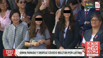 Una mujer con chaqueta gris y blusa blanca sonríe, junto a dos mujeres jóvenes con cabello oscuro y rostros cubiertos. Alrededor hay otras personas y logos de televisión