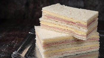 Varios sándwiches de miga con capas de jamón y queso apilados sobre una bandeja metálica plateada.