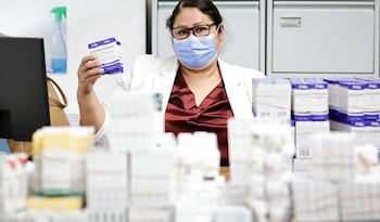 Mujer con mascarilla y lentes sostiene un paquete de cajas de paracetamol, rodeada de pilas de medicamentos en lo que parece ser una farmacia o almacén
