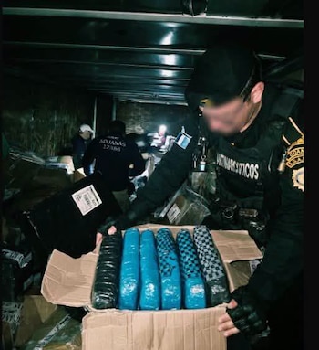 Agente con gorra y chaleco revisa caja con seis paquetes envueltos en plástico azul y estampado a cuadros. Otros agentes inspeccionan cajas