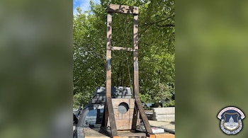 Una guillotina de madera instalada en la plataforma de una camioneta negra, con árboles al fondo y el sello de la Policía del Capitolio