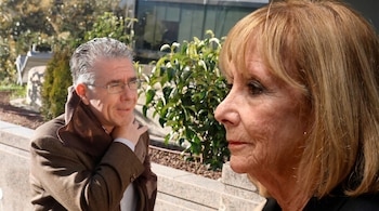 Primer plano de un hombre con cabello gris y gafas, vestido con chaqueta marrón y bufanda, junto a una mujer de perfil con cabello rubio, al aire libre.