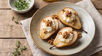 Tres mitades de pera con queso ricotta, nueces y miel en un plato de cerámica sobre una mesa de madera.
