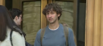 Primer plano de Misha Vitels, un joven con cabello rizado y una mochila, vistiendo una camiseta azul, parado frente a la entrada de un edificio