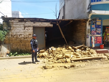 Un oficial de policía con uniforme azul de pie frente a una estructura de adobe y lámina parcialmente derrumbada. Escombros y calle adoquinada