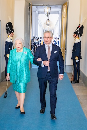 Mujer mayor de cabello blanco y traje verde azulado con un hombre en traje azul oscuro, escoltados por dos guardias uniformados en un pasillo