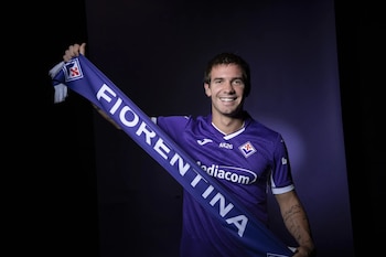 Hombre sonriente sostiene bufanda violeta con la palabra Fiorentina y un escudo. Viste camiseta violeta con logos de patrocinadores y emblema de club