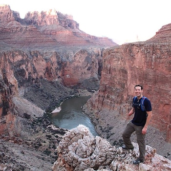 Aron Ralston sonríe, parado sobre rocas en un cañón. Viste camiseta oscura y mochila azul. Su brazo derecho amputado es visible. El río y paredes rocosas de fondo