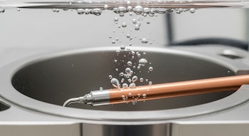 Primer plano de un reactor electroquímico con un electrodo de cobre sumergido en una solución transparente y burbujas ascendiendo a la superficie.