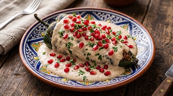 Chile poblano relleno cubierto con crema blanca, semillas de granada y perejil, sobre un plato decorado de cerámica y mesa de madera.