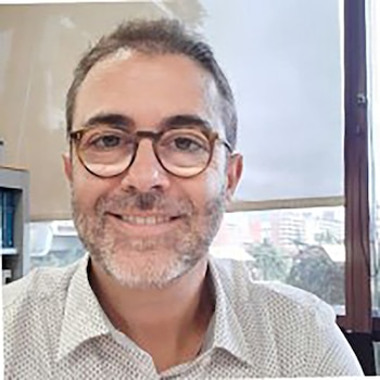 Un hombre caucásico de mediana edad, con cabello corto, barba y gafas marrones, sonríe a la cámara mientras lleva una camisa estampada, con una ventana de oficina al fondo