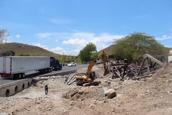 Una excavadora amarilla trabaja sobre la tierra junto a la estructura de un puente caído y una carretera con un camión